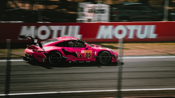 A sleek, high-performance sports car, painted in vibrant pink, speeds along a racetrack. Blurred motion lines in the background emphasize the car's speed. Advertising banners and a fenced-off area are visible along the track.