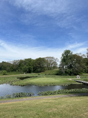 A serene landscape featuring a golf course with a green surrounded by water, set against a backdrop of lush green trees and a blue sky. A golf cart is visible near a narrow wooden bridge that crosses the water.