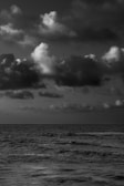A monochrome photograph of the ocean with a slightly turbulent surface beneath a dramatic sky filled with large, fluffy clouds.