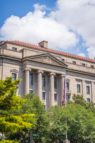 A large neoclassical building with columns and a red-tiled roof. A flagpole with the American flag is prominently displayed. Green trees are in the foreground, and a bright blue sky with fluffy white clouds is in the background.