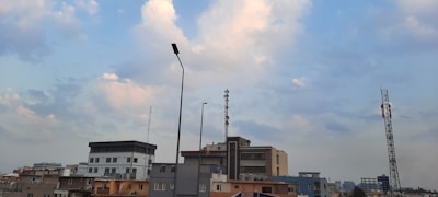 Urban skyline featuring several buildings of varying heights. There are visible telecommunications towers and streetlights protruding above the structures. The sky is mostly clear with a scattering of white and light gray clouds.