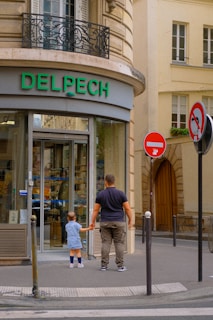 A man and a child are standing in front of a store with a glass entrance. The store has the name 'Delpech' in green letters above the door. They are holding hands, facing the entrance, in an urban setting with a beige building facade and two signs visible, including a no entry sign.