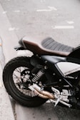A close-up view of the rear section of a motorcycle, featuring the seat, rear suspension, and exhaust system. The motorcycle is positioned on a paved surface, with visible pavement markings in the background.