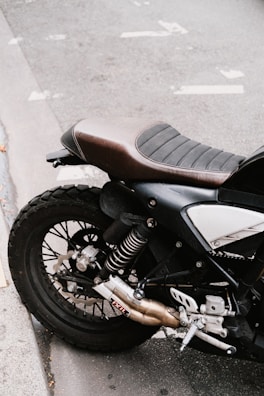A close-up view of the rear section of a motorcycle, featuring the seat, rear suspension, and exhaust system. The motorcycle is positioned on a paved surface, with visible pavement markings in the background.