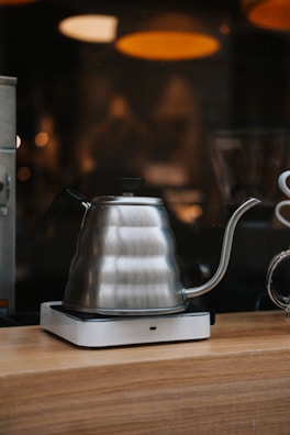 A stainless steel gooseneck kettle is positioned on a wooden surface with a soft blurred background featuring warm lighting. The kettle is placed on a small, modern heating base. The elegant curved spout and handle are prominently visible, suggesting a setting for precise coffee or tea brewing.