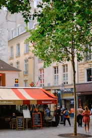 A charming city street features a small restaurant with a red and white striped awning. The outdoor seating area is adorned with menu boards. People stroll past, adding a lively atmosphere. In the background, classic European architecture with light-colored buildings can be seen, along with some green trees providing shade.