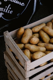 A wooden crate filled with numerous brown potatoes is positioned in front of a dark background with partial text visible.
