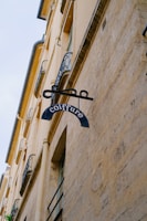 A vintage-style sign for a hair salon hangs from the side of an old building with beige stone walls. The word 'coiffure' is written on the sign, which consists of a blue metal piece attached to a decorative iron bracket. The building features ornate wrought iron railings on the windows and a classic architectural style. The overall scene is set against a pale sky.