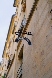 A vintage-style sign for a hair salon hangs from the side of an old building with beige stone walls. The word 'coiffure' is written on the sign, which consists of a blue metal piece attached to a decorative iron bracket. The building features ornate wrought iron railings on the windows and a classic architectural style. The overall scene is set against a pale sky.