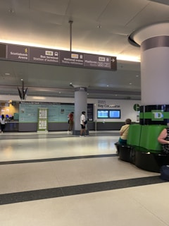 A spacious indoor transit terminal with signs indicating directions to the Scotiabank Arena, bus terminal, and national train platforms. The area has seating, informational displays, and people walking or sitting. Overhead, large signs provide navigation information, and a green column with seating surrounds it.