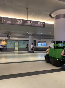 A spacious indoor transit terminal with signs indicating directions to the Scotiabank Arena, bus terminal, and national train platforms. The area has seating, informational displays, and people walking or sitting. Overhead, large signs provide navigation information, and a green column with seating surrounds it.