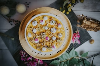 A beautifully decorated cake with an ornate pattern of flowers and icing on top. The cake is presented on a circular wooden tray, surrounded by a rustic setting that includes a decorative deer figure, some floral accents, and greenery.