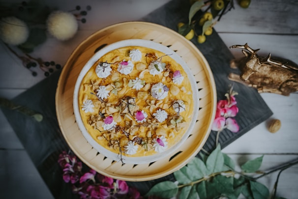 A beautifully decorated cake with an ornate pattern of flowers and icing on top. The cake is presented on a circular wooden tray, surrounded by a rustic setting that includes a decorative deer figure, some floral accents, and greenery.