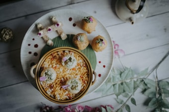 A beautifully arranged plate features a variety of snacks, including a bowl of puffed rice garnished with decorative flowers. Surrounding the bowl on the plate are small, round pastries and rolls, each topped with small floral garnishes. The setting includes subtle decorative elements like a teapot and soft floral arrangements, providing a serene and elegant dining atmosphere.