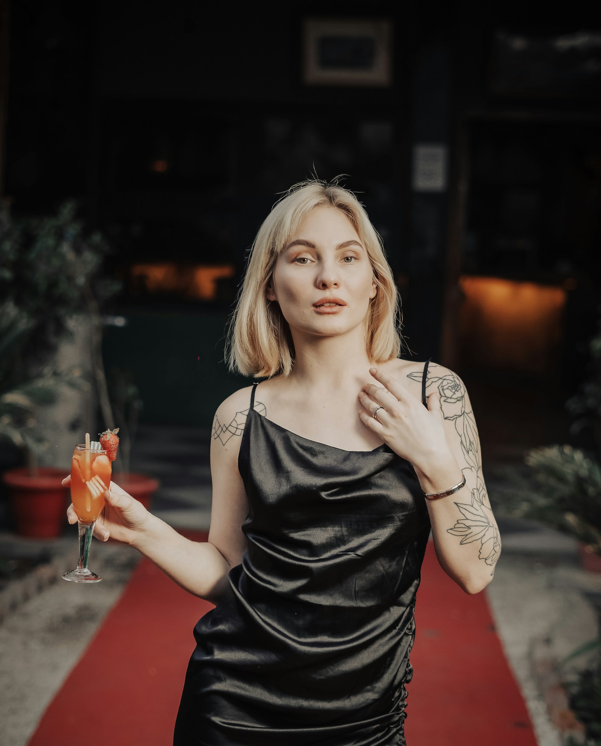 A woman with blonde hair and tattoos on her arms is wearing a black dress and holding a cocktail glass with a colorful drink, garnished with a strawberry. She stands on a red carpet, with plants and a dimly lit background.