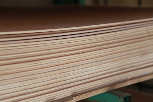 A close-up view of a stack of smooth, evenly cut plywood sheets. The wood grain and layers are visible, indicating high-quality, processed lumber. The sheets are neatly aligned and resting on a green support structure.