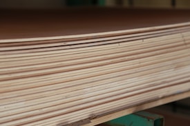 A close-up view of a stack of smooth, evenly cut plywood sheets. The wood grain and layers are visible, indicating high-quality, processed lumber. The sheets are neatly aligned and resting on a green support structure.