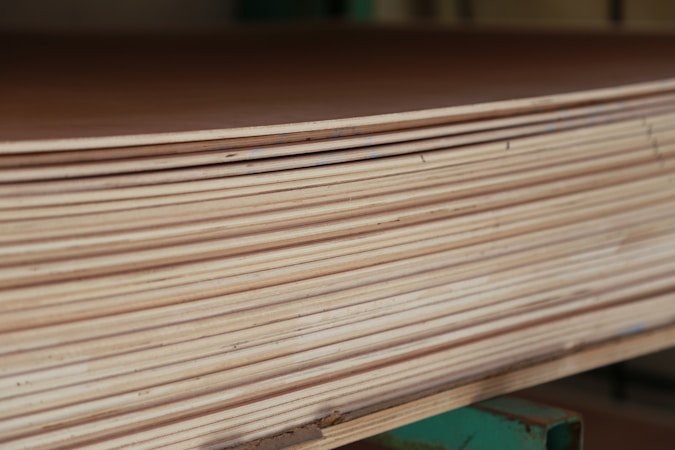 A close-up view of a stack of smooth, evenly cut plywood sheets. The wood grain and layers are visible, indicating high-quality, processed lumber. The sheets are neatly aligned and resting on a green support structure.