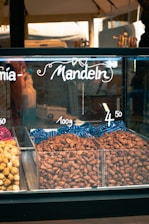 A display case filled with nuts labeled as 'Mandeln' indicating almonds. The almonds are arranged in plastic containers, with pricing displayed above them. The setting appears to be a market or a store.