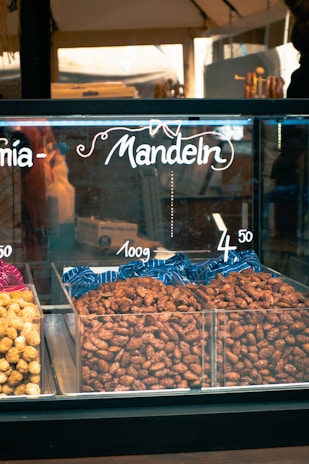 A display case filled with nuts labeled as 'Mandeln' indicating almonds. The almonds are arranged in plastic containers, with pricing displayed above them. The setting appears to be a market or a store.