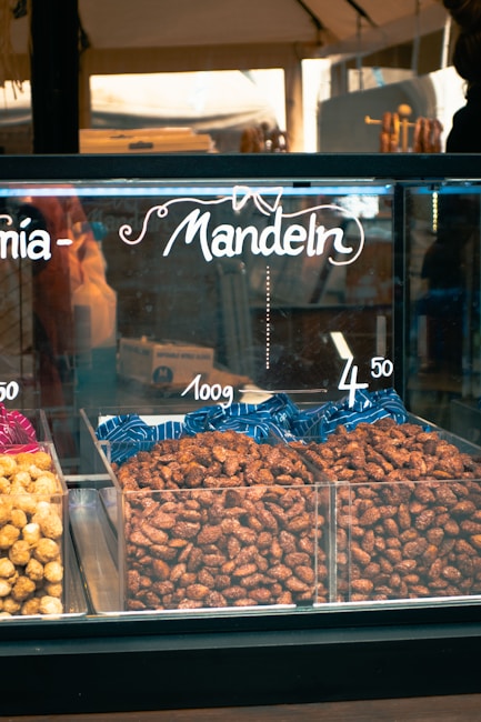 A display case filled with nuts labeled as 'Mandeln' indicating almonds. The almonds are arranged in plastic containers, with pricing displayed above them. The setting appears to be a market or a store.