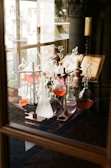 A collection of glass laboratory equipment with various colored liquids is arranged on a wooden table. An open book with diagrams is placed nearby. The setting appears to be a vintage or historical laboratory with dim lighting and wooden elements.