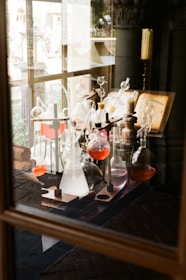 A collection of glass laboratory equipment with various colored liquids is arranged on a wooden table. An open book with diagrams is placed nearby. The setting appears to be a vintage or historical laboratory with dim lighting and wooden elements.