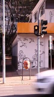 A street scene featuring a building with an artwork painted on the wall, showing a man playing a guitar and a woman sitting next to him. Above them are illustrations of birds flying. A traffic light with a red signal is in the foreground, alongside a sidewalk with a trash bin. Trees and an urban apartment building form the background.