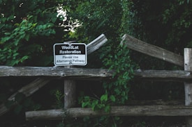 A wooden fence surrounded by lush green foliage, featuring a sign that reads 'WoodLot Restoration. Please Use Alternative Pathway.' The scene appears to be in a natural, wooded area with dense vegetation.