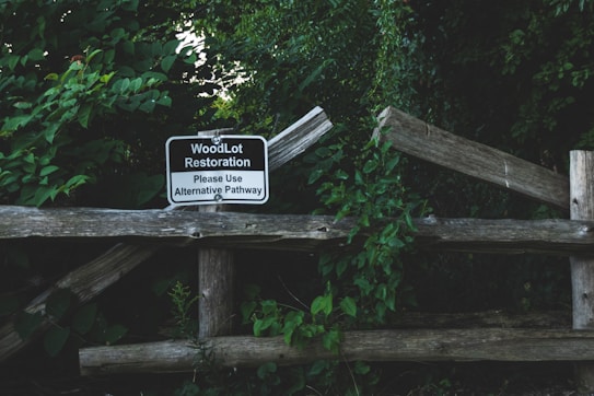A wooden fence surrounded by lush green foliage, featuring a sign that reads 'WoodLot Restoration. Please Use Alternative Pathway.' The scene appears to be in a natural, wooded area with dense vegetation.