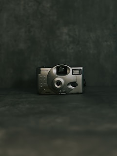 A vintage-style camera with a metallic finish is centered against a dark, textured background. The camera features a circular lens and simple controls, suggesting a minimalist, retro design.