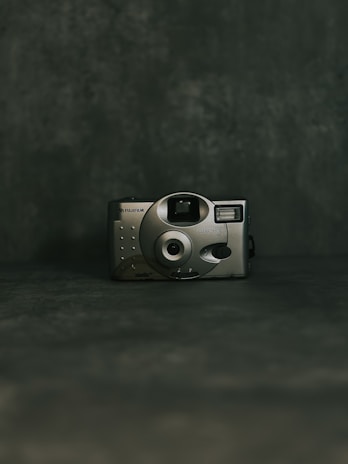 A vintage-style camera with a metallic finish is centered against a dark, textured background. The camera features a circular lens and simple controls, suggesting a minimalist, retro design.