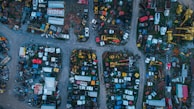 Aerial view of an extensive junkyard with various old and abandoned vehicles, including cars, trucks, and trailers. The area is divided into multiple sections by dirt paths, and various metal parts and machinery are scattered throughout. The vehicles and parts are in different stages of disrepair, with rust and wear evident.