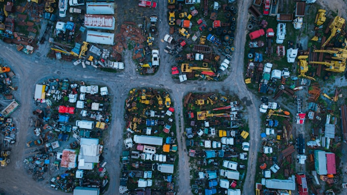 Aerial view of an extensive junkyard with various old and abandoned vehicles, including cars, trucks, and trailers. The area is divided into multiple sections by dirt paths, and various metal parts and machinery are scattered throughout. The vehicles and parts are in different stages of disrepair, with rust and wear evident.