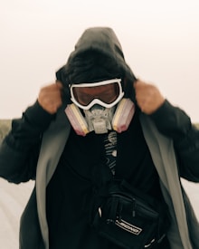 A person is wearing a black hooded jacket and a protective mask with filters, adjusting the hood over their head. They are also wearing ski goggles and have a black crossbody bag. The background appears to be a muted outdoor setting, possibly sandy or desert-like.