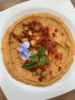 A plate of hummus garnished with whole chickpeas, diced red pepper, a sprig of mint, and a small blue flower. The hummus is sprinkled with paprika and is served on a white plate placed on a wooden surface.