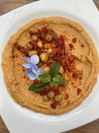 A plate of hummus garnished with whole chickpeas, diced red pepper, a sprig of mint, and a small blue flower. The hummus is sprinkled with paprika and is served on a white plate placed on a wooden surface.