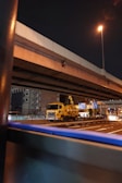 A nighttime urban scene featuring an overpass with a yellow utility truck positioned underneath. Streetlights illuminate the area, casting a warm glow over the road. Buildings and infrastructure with directional signs are visible in the background.
