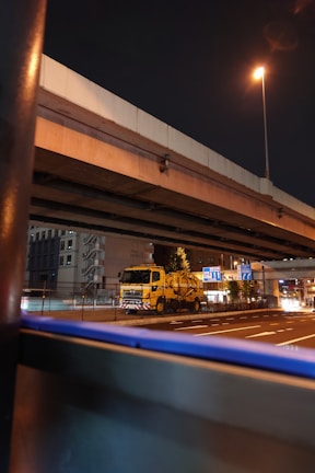 A nighttime urban scene featuring an overpass with a yellow utility truck positioned underneath. Streetlights illuminate the area, casting a warm glow over the road. Buildings and infrastructure with directional signs are visible in the background.