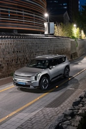 A modern silver SUV parked on a narrow, stone-lined street. The street is dimly lit by overhead lamps, and a sleek, contemporary building with horizontal stripes is visible on the left. The scene conveys an urban and sophisticated ambiance with elements of greenery and traditional architecture in the background.