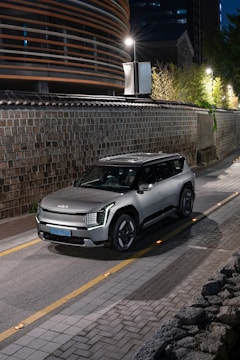 A modern silver SUV parked on a narrow, stone-lined street. The street is dimly lit by overhead lamps, and a sleek, contemporary building with horizontal stripes is visible on the left. The scene conveys an urban and sophisticated ambiance with elements of greenery and traditional architecture in the background.
