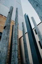 Tall wooden poles with inscriptions are positioned vertically against the backdrop of a modern building. The poles are weathered, adding a rustic feel, while the building features a mix of concrete and glass. The perspective is from below, looking upwards.