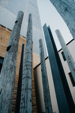 Tall wooden poles with inscriptions are positioned vertically against the backdrop of a modern building. The poles are weathered, adding a rustic feel, while the building features a mix of concrete and glass. The perspective is from below, looking upwards.