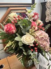 A vibrant floral arrangement featuring a mix of roses, hydrangeas, and various greenery arranged in a glass vase. The colors range from white to deep pink and red, accented by small red berries. The background shows a wooden frame and some crochet fabric.