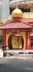 A small, colorful mosque entrance with a prominent yellow and red color scheme. The mosque features a golden dome at the top and is located next to a street, with a white car and a parked motorcycle in front. The architecture includes an arched entrance with decorative pillars and a sign that reads 'Masjid Al-Imtizaj'.