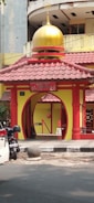A small, colorful mosque entrance with a prominent yellow and red color scheme. The mosque features a golden dome at the top and is located next to a street, with a white car and a parked motorcycle in front. The architecture includes an arched entrance with decorative pillars and a sign that reads 'Masjid Al-Imtizaj'.