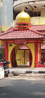 A small, colorful mosque entrance with a prominent yellow and red color scheme. The mosque features a golden dome at the top and is located next to a street, with a white car and a parked motorcycle in front. The architecture includes an arched entrance with decorative pillars and a sign that reads 'Masjid Al-Imtizaj'.
