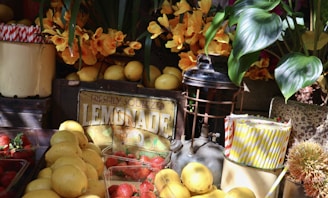 A vibrant market stall featuring a display of fresh lemons and strawberries arranged around a vintage-style metal sign with the word 'LEMONADE'. The scene includes yellow and white striped straws in a container and is decorated with lush green leaves and bright yellow flowers.