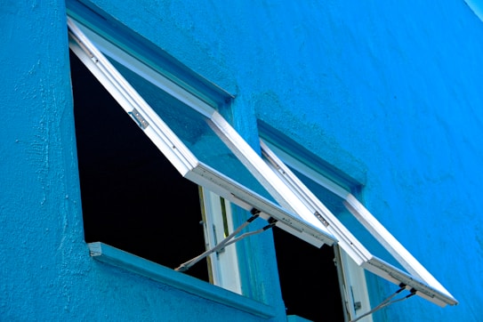 Two open windows are set in a vibrant blue wall. The windows are hinged at the top and are opened outward. The wall appears to have a textured surface, with the blue paint giving a fresh and clean look.