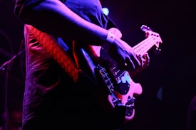 A close-up view of a person playing an electric guitar, focusing on their hands and the strings. The scene is dimly lit, with vibrant purple and blue lighting creating a dramatic atmosphere. The musician's red strap and the glossy finish of the guitar stand out.